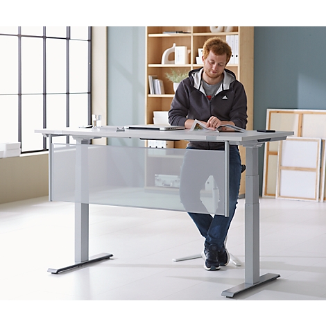 A man with a beard leans on a standing desk, reading a book. He wears a jacket and jeans. In the background: window, shelf, paintings.