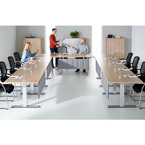 Salle de conférence avec des meubles clairs. Trois personnes sont assises à une table en forme de U, une personne remet des papiers.