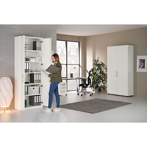 A woman stands in an office at a white cabinet, holding files, looking inside.