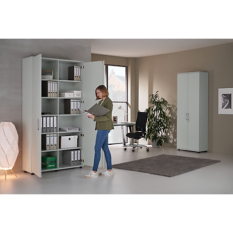 A woman stands in front of an open filing cabinet, looking at documents. In the background, another cabinet, desk, and a window.