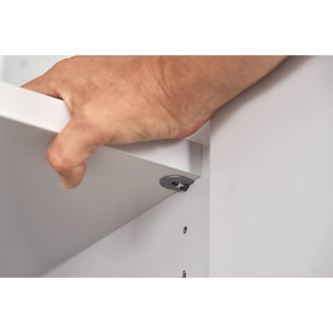 A hand holds a white shelf, which is attached to a gray holder. In the background a white wall.