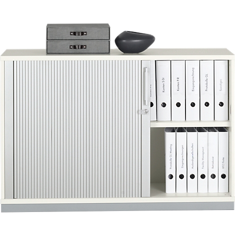 White cabinet with roller shutter, grey drawers and black vase. Open compartments with white binders.