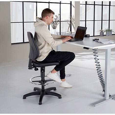 Un homme est assis à un bureau réglable en hauteur, travaillant sur un ordinateur portable. Il est assis sur une chaise pivotante noire avec repose-pieds. Le bureau est lumineux.