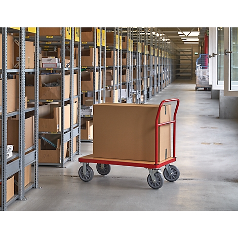 Un carton sur un chariot rouge dans un entrepôt. Rayonnages de boîtes à gauche, allées en arrière-plan.