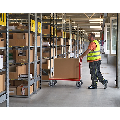 Un homme portant un gilet de sécurité pousse un chariot avec une boîte en carton dans un entrepôt. Étagères avec boîtes, panneaux jaunes avec des numéros.