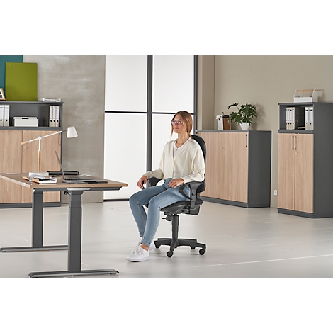 Woman seated in an office chair at a height-adjustable desk. Office furniture in the background.