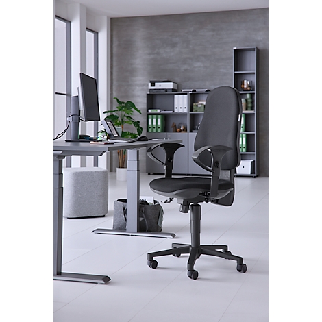 Office chair in front of a gray background. Desk, screen, plants, windows and shelves in the background. Light gray floor.