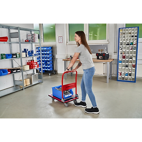 A woman pulls a red cart with a blue crate in a workshop. In the background, shelves with materials.