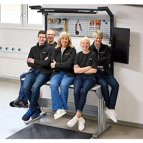 Vijf mensen zitten op een werkbank. Achter hen een pegboard met gereedschap, een monitor.