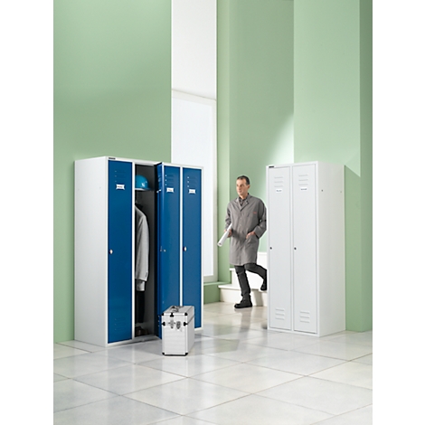 A man walks past lockers in a lab. One locker is open. A blue cap, a coat and a toolbox are ready.