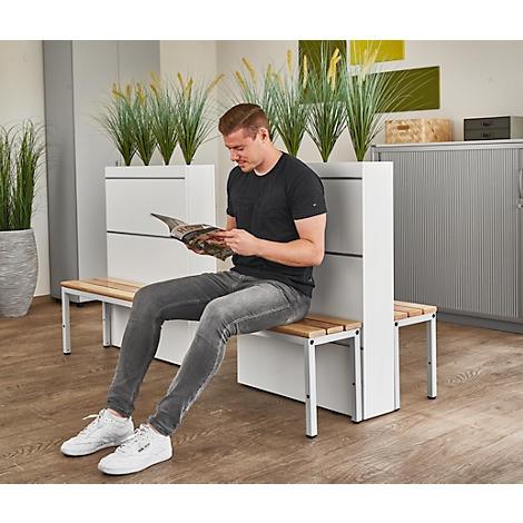 A man sits on a bench reading a magazine in a waiting area. Plants are behind him.
