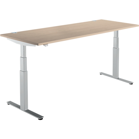 Un bureau réglable en hauteur avec un plateau de table brun clair et des pieds argentés. Le bureau se dresse sur un fond blanc.
