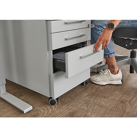 Une personne ouvre le tiroir d'un caisson blanc. Parquet en bois au sol. Tatouage et montre sur la jambe.