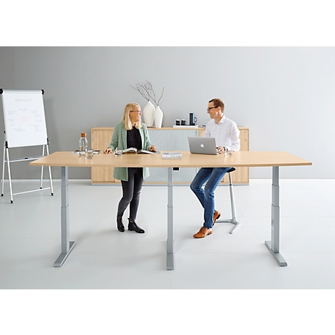 Two people at a height-adjustable desk, a woman standing, the man sitting, whiteboard and sideboard in the background.