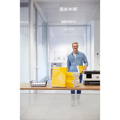 A man stands in the office. Stacks of paper and a printer are on a table.