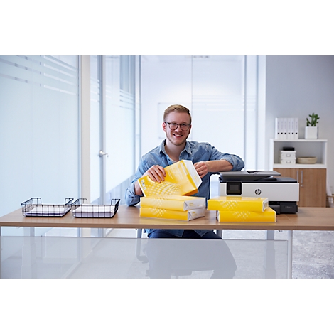 A man with glasses opens a paper package at his desk. A printer stands on the table.