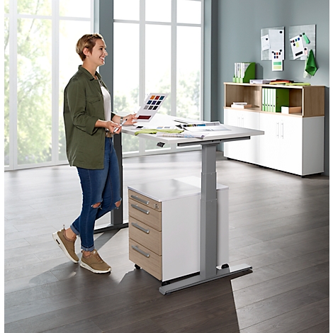 A woman stands at a standing desk, looking at color samples. She wears jeans and a jacket. The desk is light, the background gray.