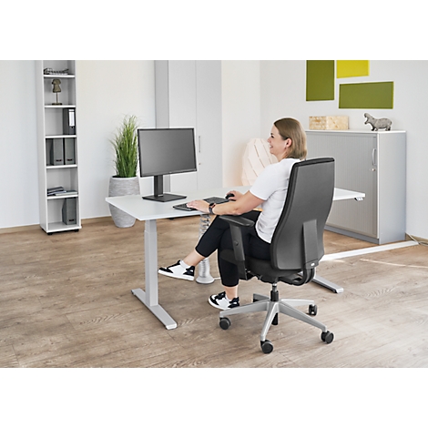 Woman sitting at a desk working on the computer. Grey desk, black office chair, white wall.