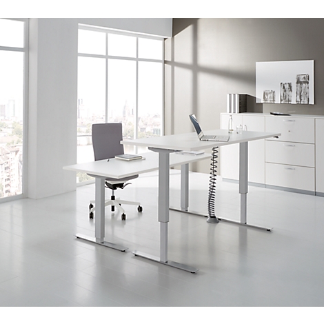 Un espacio de oficina luminoso con un escritorio ajustable en altura, una silla y ventanas con vistas a la ciudad.