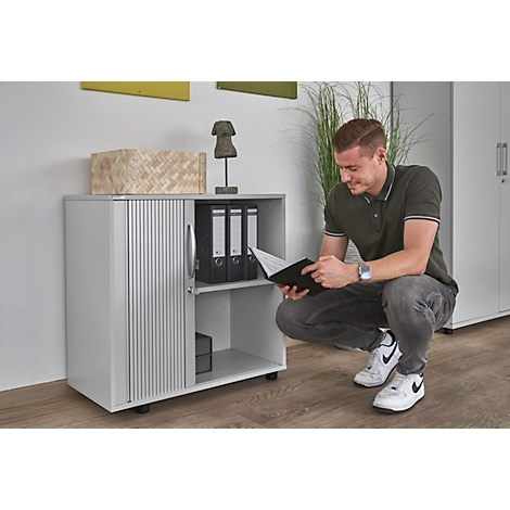 Man squats by a gray cabinet, reading a book. The cabinet has a roller shutter. File folders sit on the shelf.
