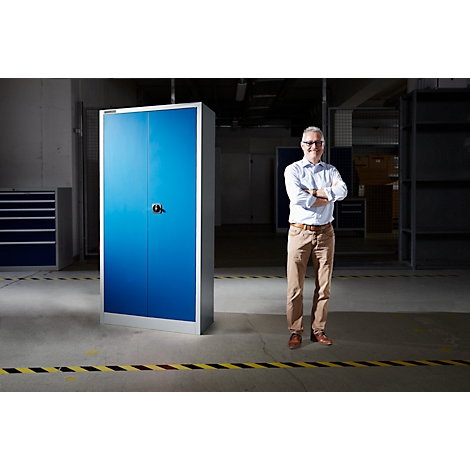 Man in magazijn voor blauwe kast. Hij draagt overhemd, broek, bril en gekruiste armen.