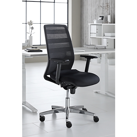 Black office chair with mesh back in front of light background. The chair has armrests and castors, in the background windows and desk.