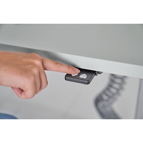 Ein Finger drückt auf eine Taste an einer Steuerung unter einem Schreibtisch. Helle, neutrale Farben.