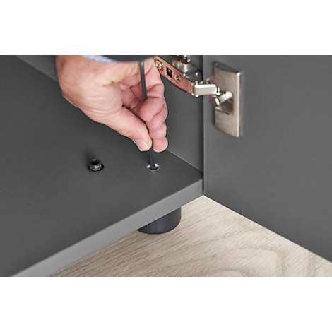Hand using an Allen key to screw a screw into a gray piece of furniture. A hinge can be seen in the background.