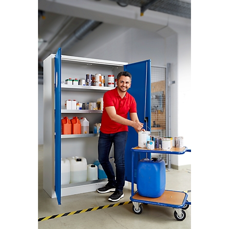 Ein Mann steht vor einem geöffneten Stahlschrank mit Farbeimern und Kanistern, lächelnd in rotem Shirt. Daneben ein Wagen.