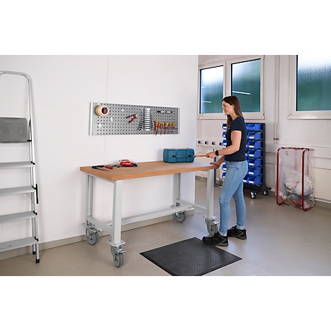 Woman standing at a workbench. Tools hang on a pegboard, next to a ladder. Blue toolbox, black mat.