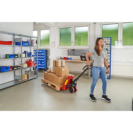 Une femme tire un transpalette avec des cartons dans un atelier. Étagères et fenêtres en arrière-plan.