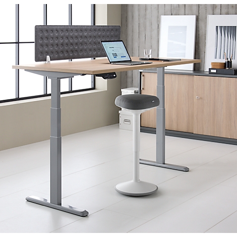 Heller Schreibtisch mit höhenverstellbarem Gestell, Laptop und Hocker; im Hintergrund graue Wand.