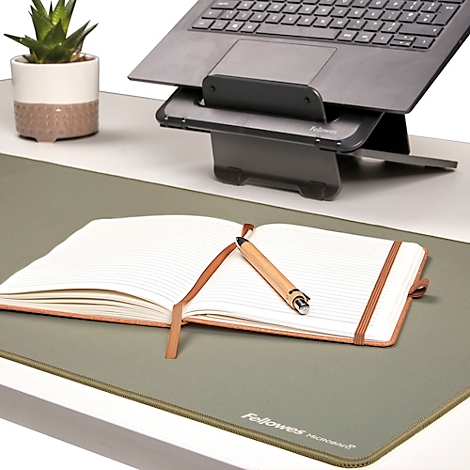 Schwarzer Laptop auf Ständer, darunter Tastatur, Maus, Notizbuch mit Stift und Pflanze. Vor weißem Hintergrund.