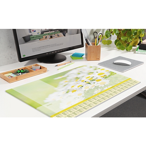 Schreibtisch mit Mauspad, Kalender und Zubehör. Im Hintergrund ein Monitor und eine Pflanze.