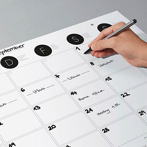 Person beschreibt mit Stift Termine in einem grossen weissen Kalender. Datum, Urlaub, Werkstatt, Home Office und Meeting sind notiert.