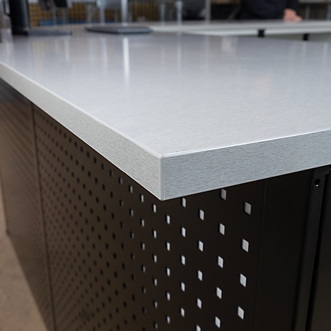 Nahaufnahme einer Schreibtischkante aus hellem Material. Der Schreibtisch darunter hat ein Lochmuster aus quadratischen Ausschnitten.