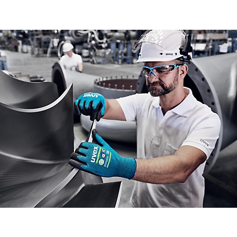 Ein Mann in Arbeitskleidung prüft ein metallenes Bauteil. Er trägt blaue Handschuhe, eine Schutzbrille und einen Helm.