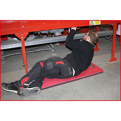 Ein Mann liegt auf einer roten Matte unter einer roten Maschine und repariert sie. Er trägt eine schwarze Hose und ein schwarzes Oberteil.