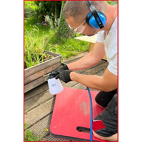 Ein Mann mit Gehörschutz und Brille kniet draußen und besprüht ein Holzkonstrukt mit einer Sprühflasche.