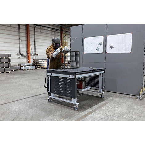 Schweißer mit Schutzhelm an Schweißtisch in Fabrik. Drahtgeflecht wird bearbeitet. Graue Wand mit Zeichnungen im Hintergrund.