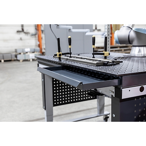 Grauer Werkzeugtisch mit Lochplatte, Schublade, Werkstücken und Schraubzwingen. Im Hintergrund ein Roboterarm.