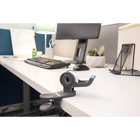 A clip-on tape dispenser on a desk. Background includes a computer, keyboard, desk lamp, and office accessories.