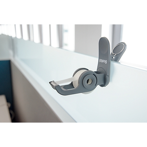 A gray scotch tape dispenser sticks to a white shelf. Transparent tape is being pulled.