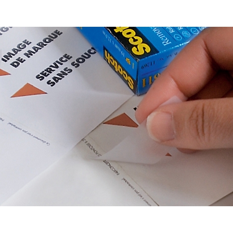 Een hand trekt voorzichtig een label van een vel papier, daarnaast ligt een blauwe Scotch-doos.