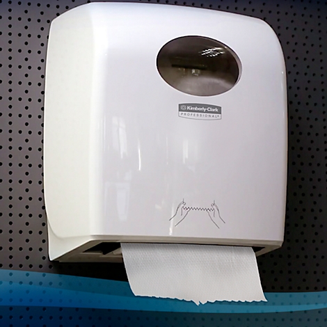 Weisser Papierhandtuchspender an einer Wand. Ein Papierhandtuch hängt heraus. Auf dem Spender steht "Kimberly-Clark Professional".