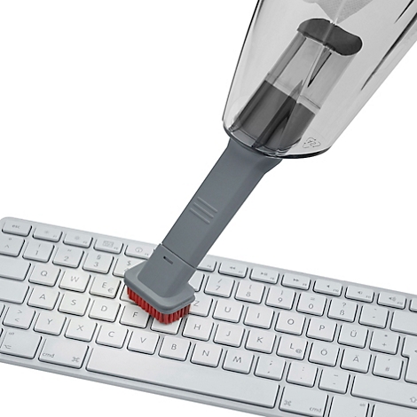 Eine Nahaufnahme zeigt einen grauen Mini-Staubsauger, der eine weiße Tastatur absaugt. Die Borsten sind rot.