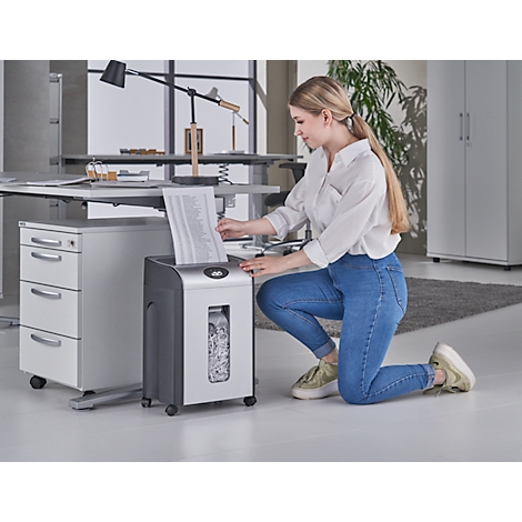 A woman kneels and puts paper into a shredder. She wears jeans and a white shirt. The room is an office.