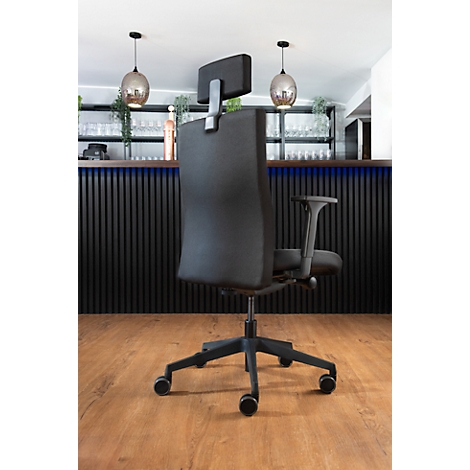 Un fauteuil de bureau noir se dresse devant un fond sombre. La chaise a un appui-tête et des roulettes.