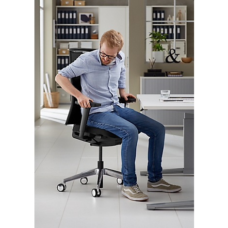 Un homme avec des lunettes est assis sur une chaise de bureau noire, en train d'ajuster les accoudoirs. Il porte un jean et une chemise bleu clair.