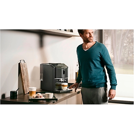 Ein Mann steht neben einer Kaffeemaschine. Auf einem Tablett stehen zwei Tassen Kaffee und Gebäck. Ein Fenster im Hintergrund.